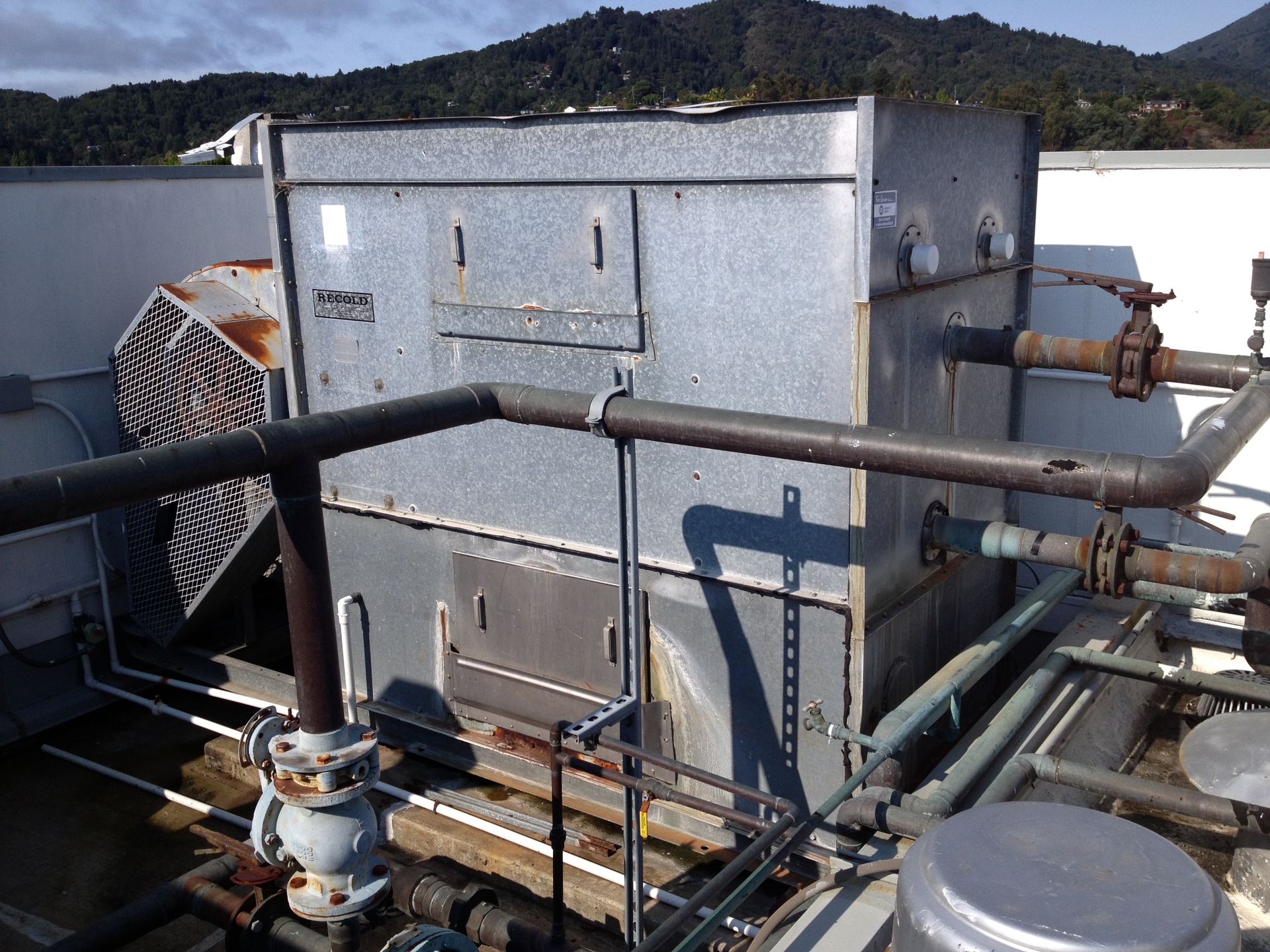 Industrial rooftop HVAC unit with pipes and railings beside a large vent fan, mountains in the background