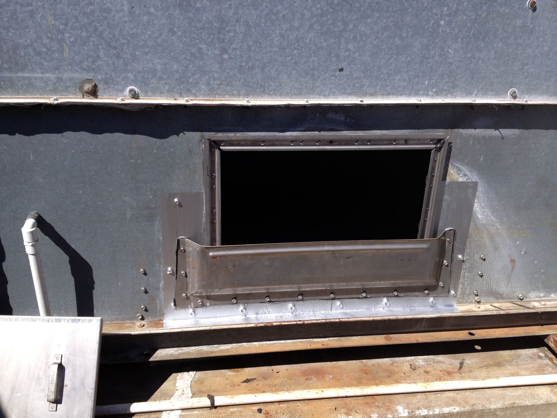 Small rectangular opening in a gray metal wall above wooden slats, with a hinged flap open below it