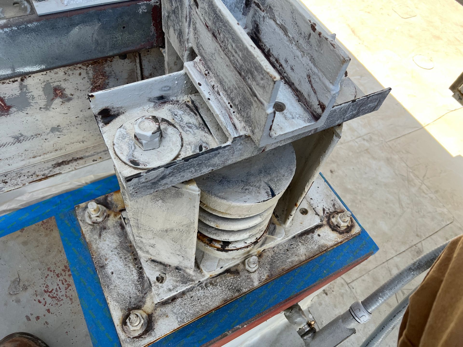 Close-up of a worn metal industrial machine base with bolts, hoses, and a blue support frame.
