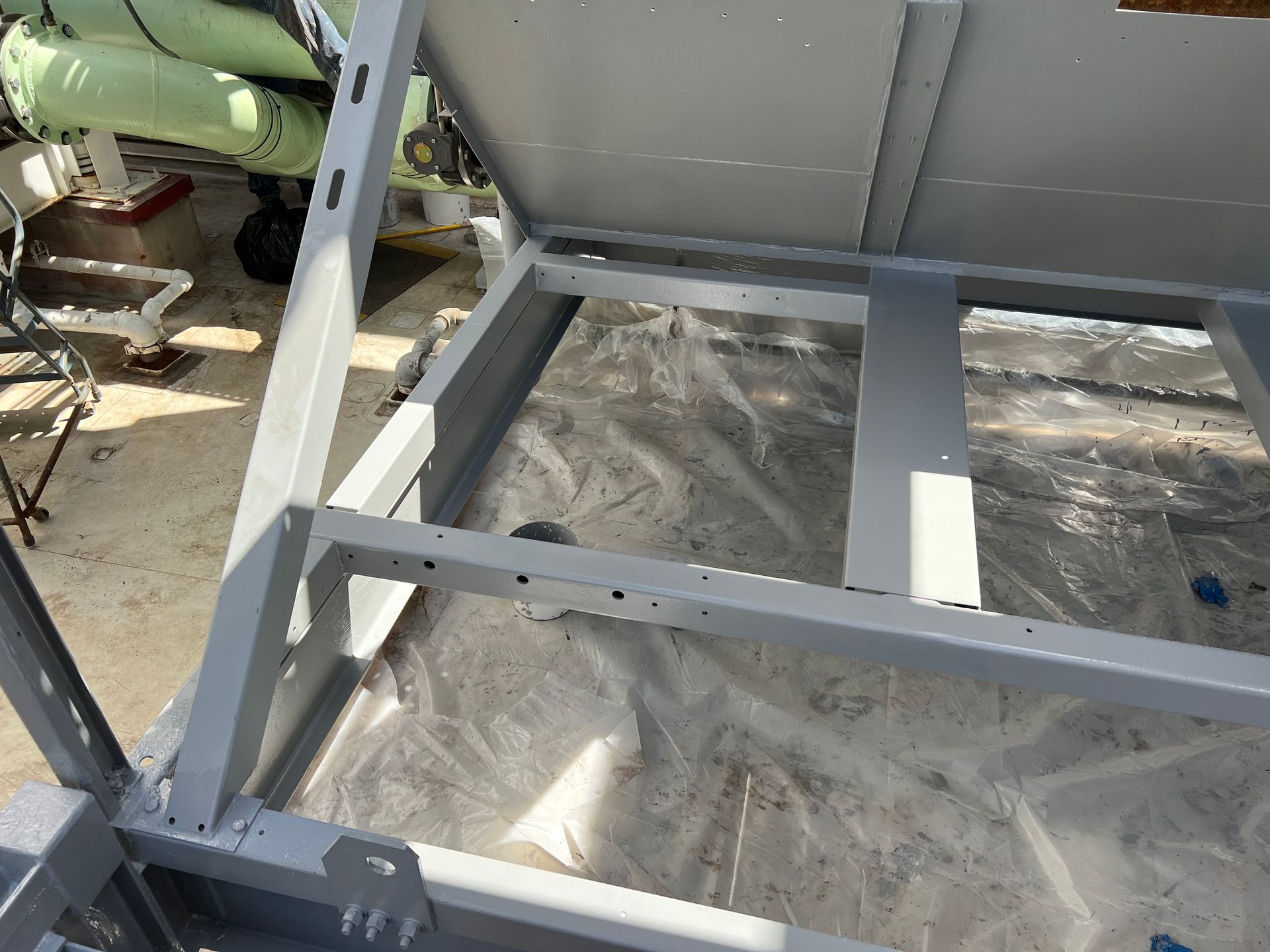 Gray metal frame structure on plastic sheeting outdoors, with crossbars and a hinged panel.