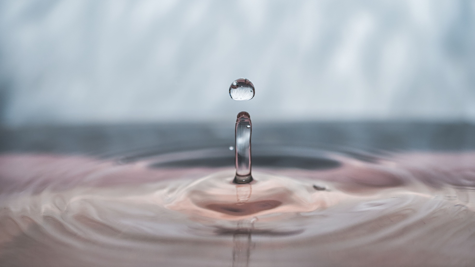 Water droplet splashing into a calm pink-tinted surface, creating a rippling crown.