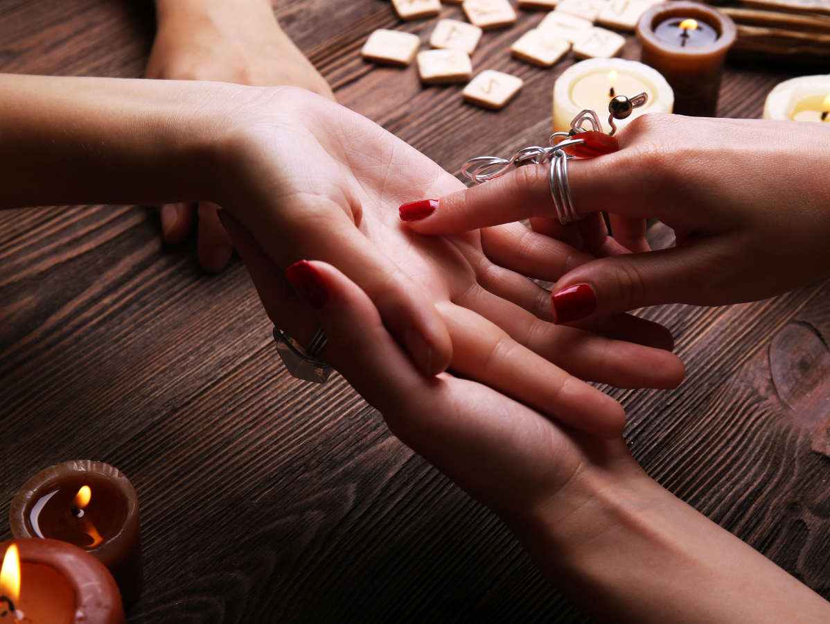 A close-up of a person reading a palm. A finger traces the lines of an open hand atop a wooden table with runes and candles.
