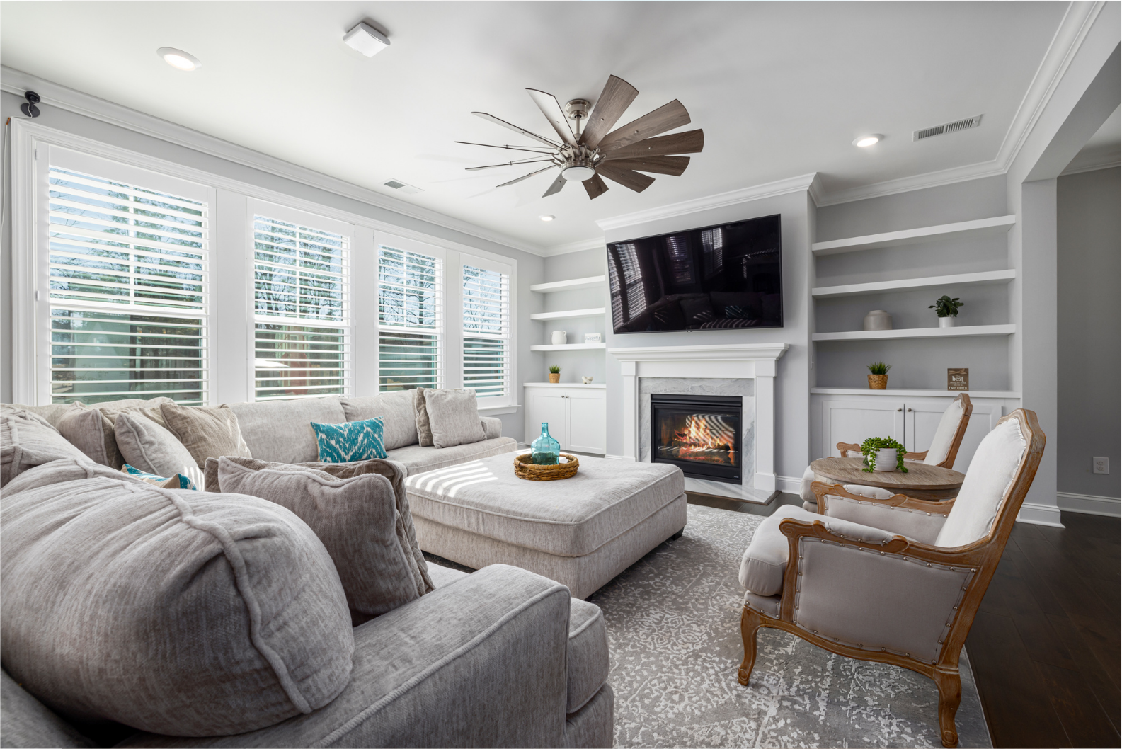 A cozy and modern living room with a large gray sectional sofa, a plush ottoman, elegant beige armchairs and ceiling fan.