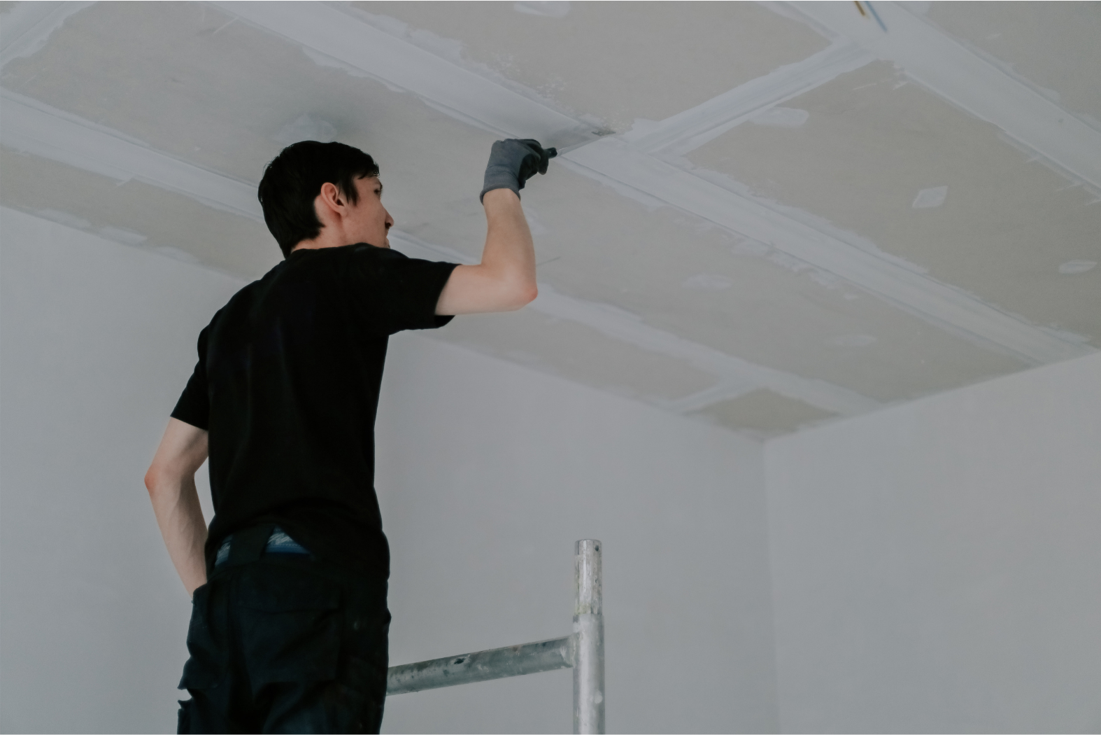 A person on a scaffold applies sealant to ceiling seams in a room