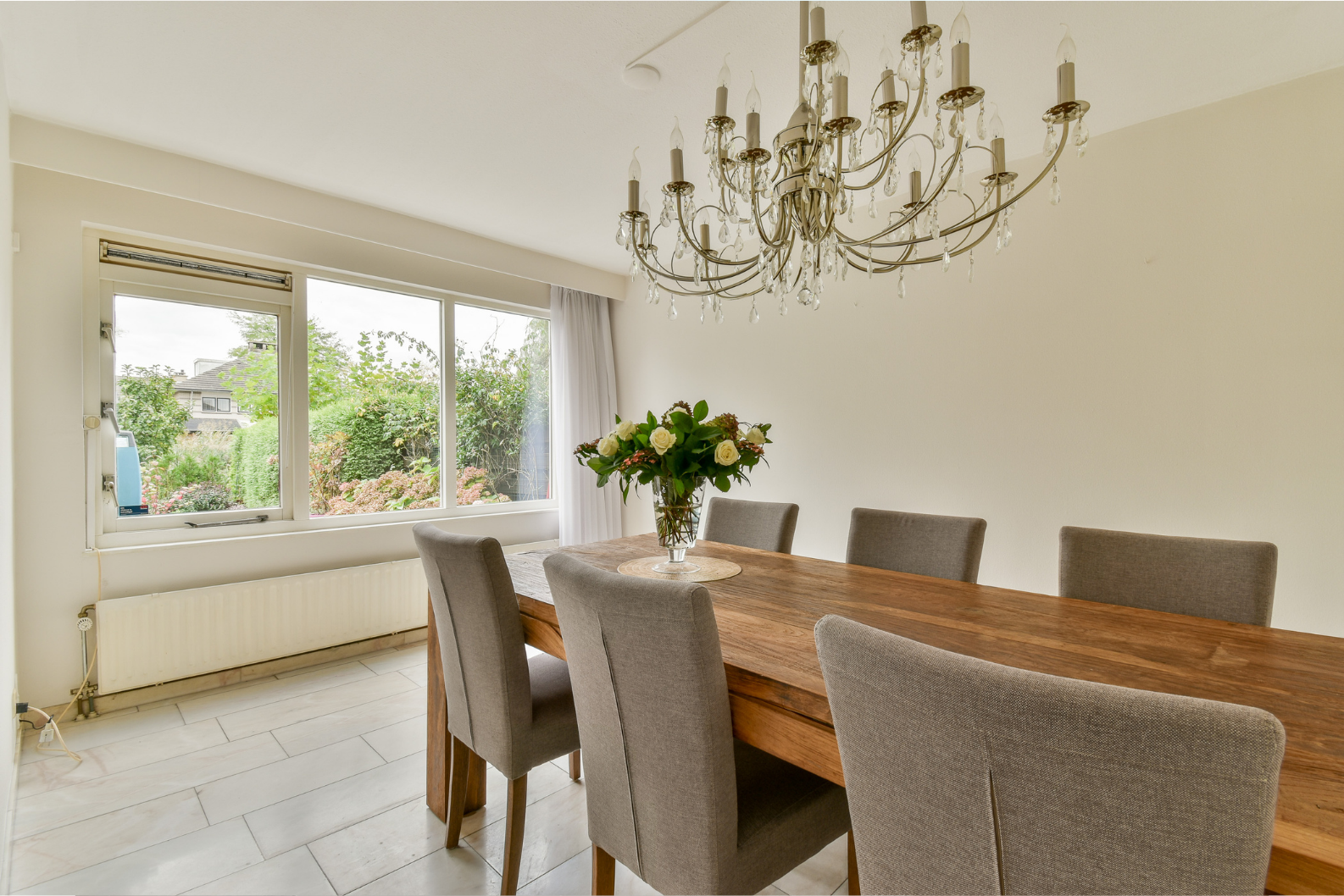Bright dining room with a wooden table, gray chairs, and a vase of flowers with elegant chandelier overhead.