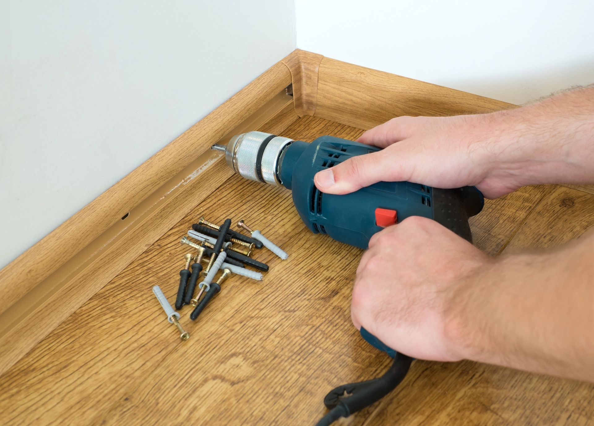 Hands using a blue drill to fasten screws in a wooden corner with scattered screws nearby.