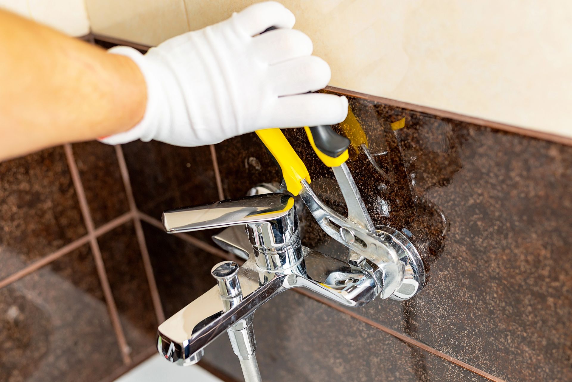 Gloved hand using pliers to repair a chrome faucet in a tiled bathroom sink area