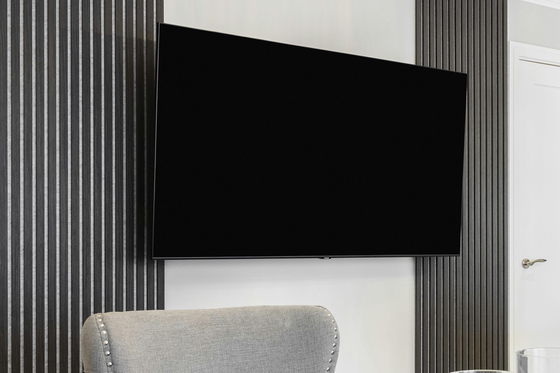 A large, black flat-screen TV mounted on a gray and white striped wall, above a gray upholstered chair.