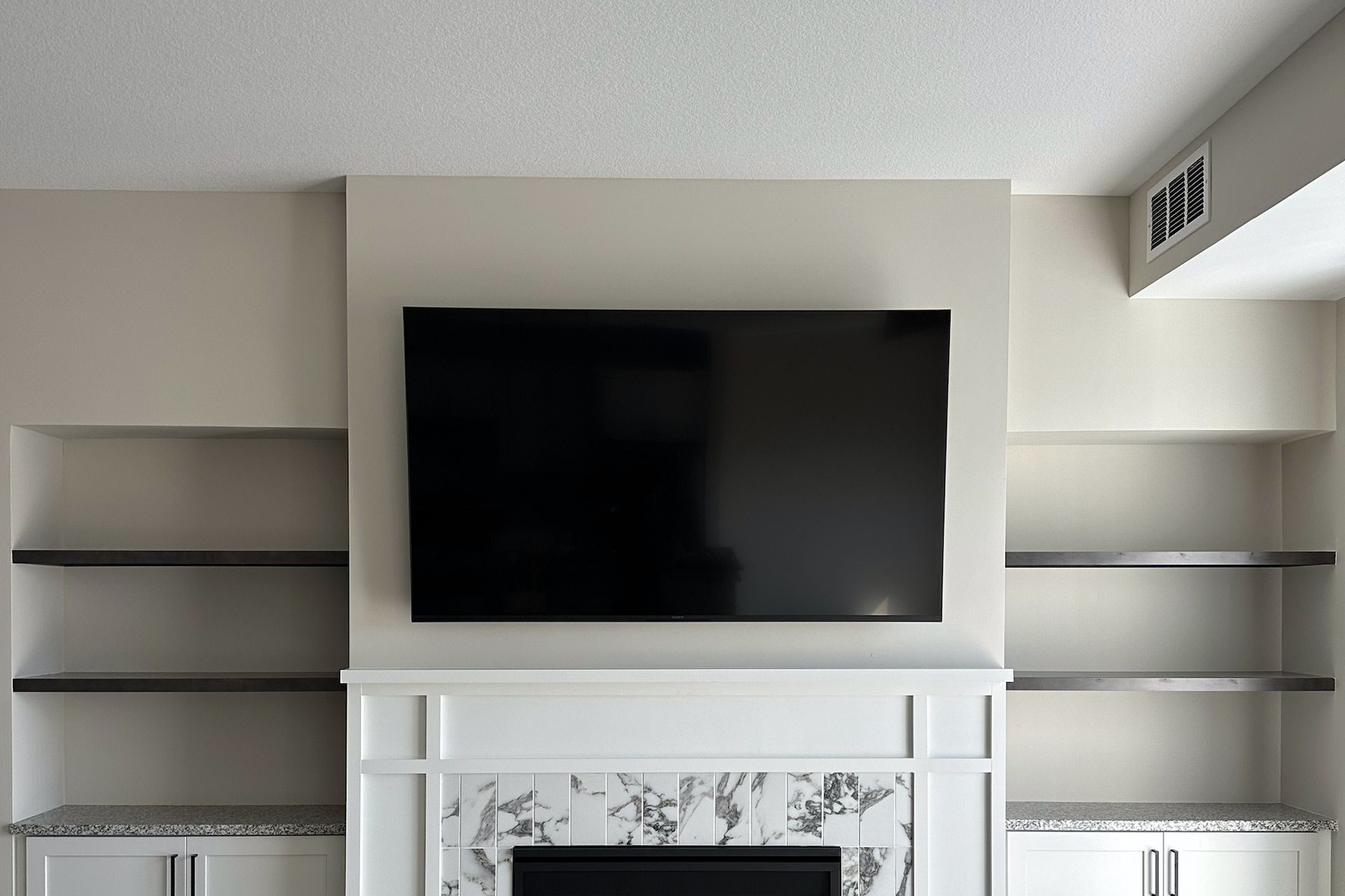 Large TV above a white fireplace, flanked by built-in shelves. Beige wall, bright room.