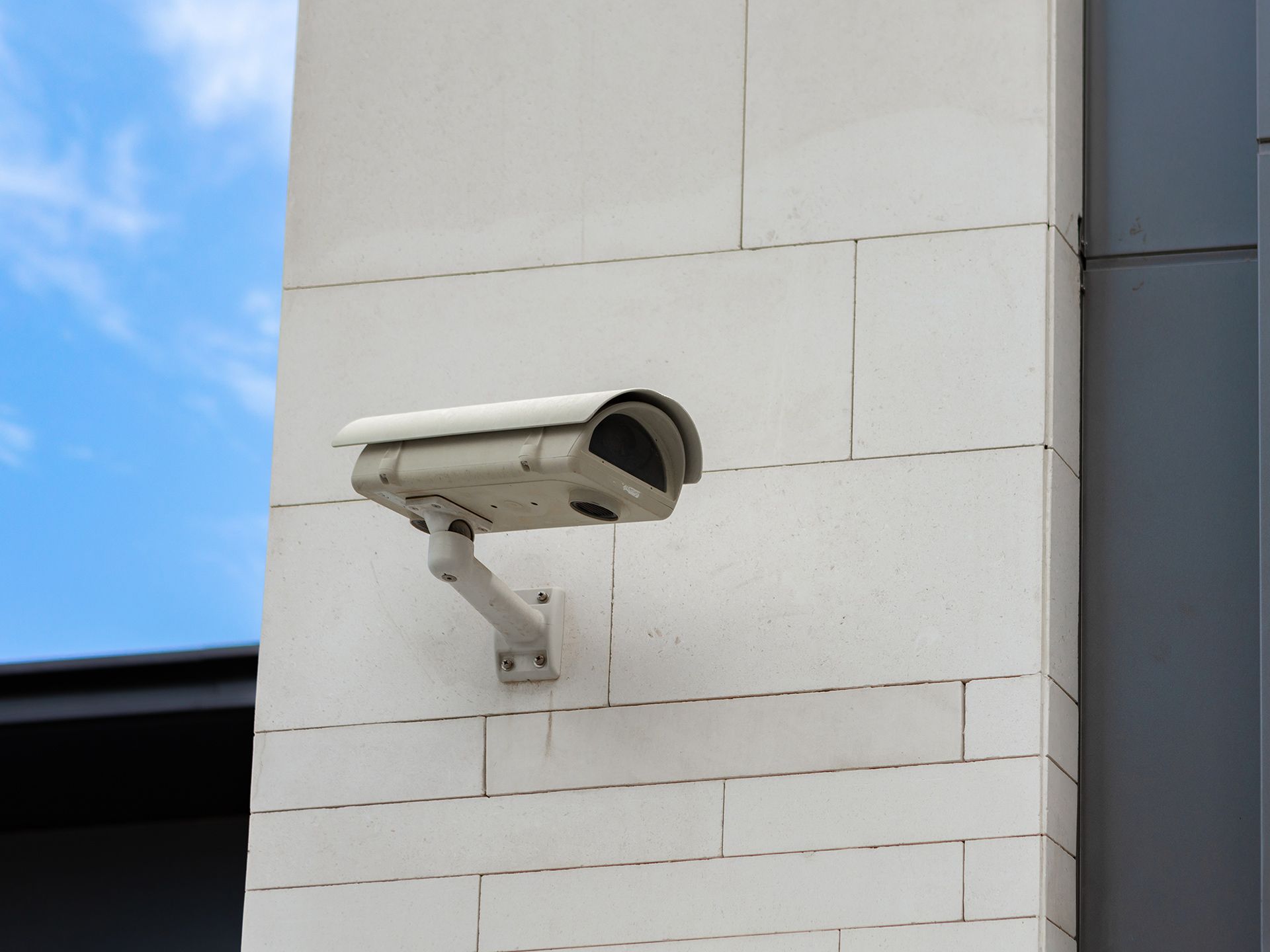 Security camera mounted on a white brick building, angled towards an unseen area; blue sky in the background.
