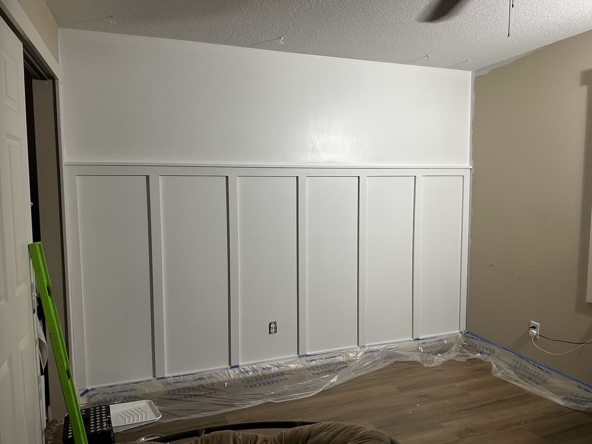 White paneled accent wall being painted, room covered in plastic, door on left.