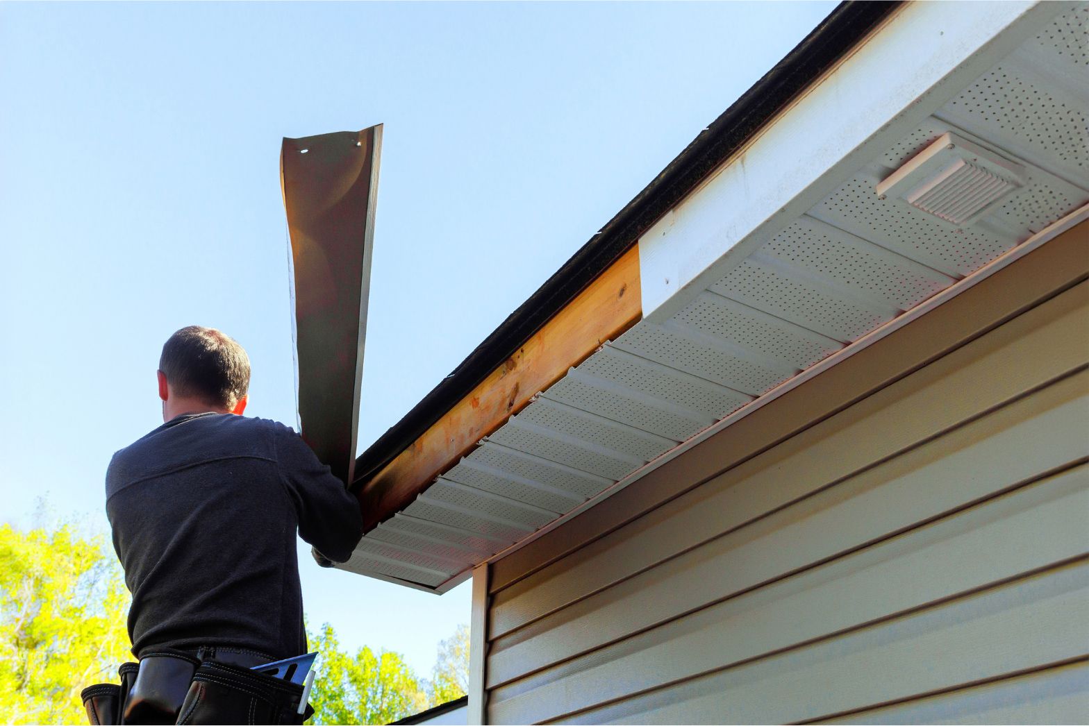 Person installing a gutter on a house roofline with a ladder nearby