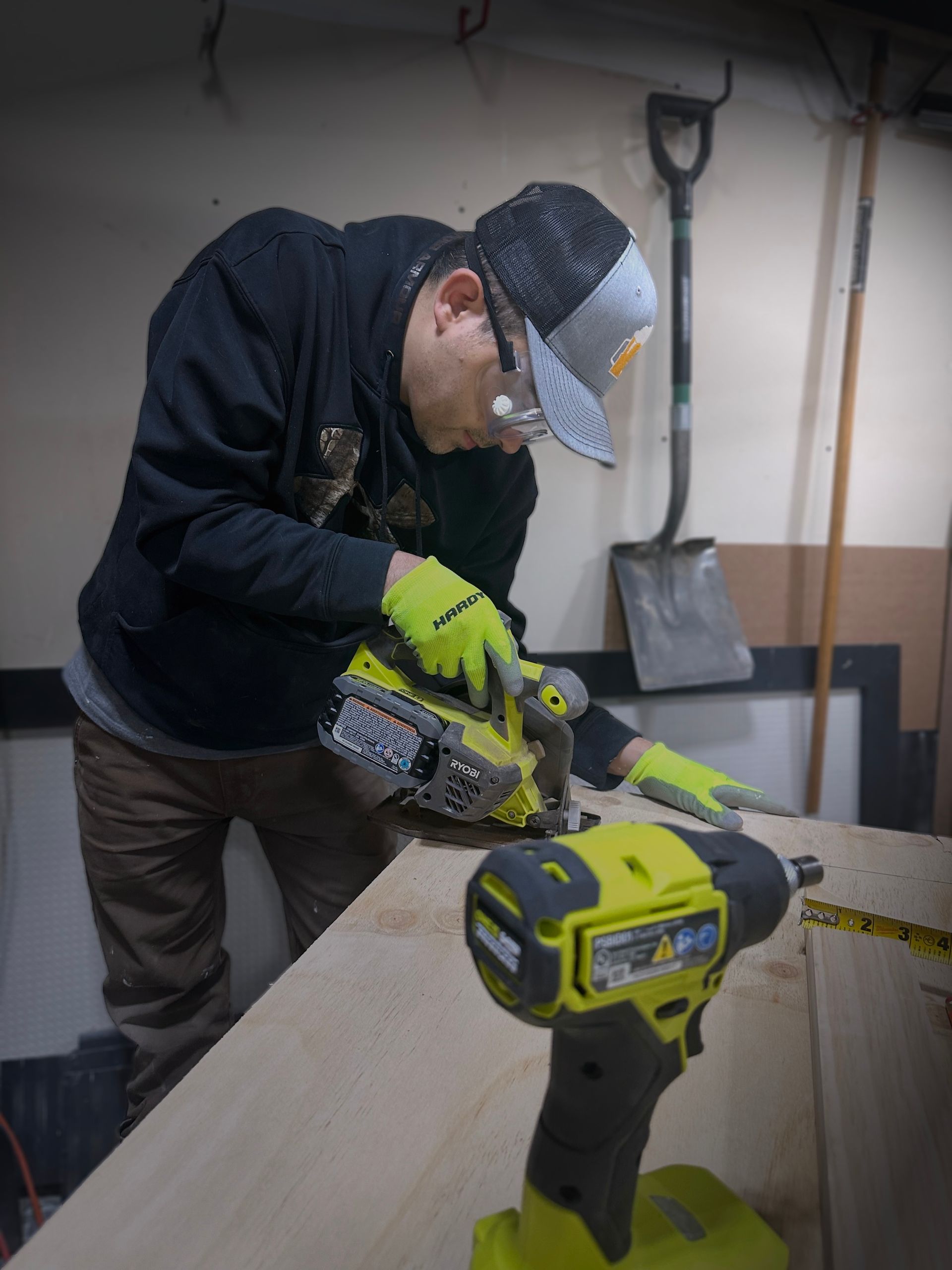 Man using a power tool to cut wood; wearing safety glasses and gloves in a workshop with a second tool in the foreground.