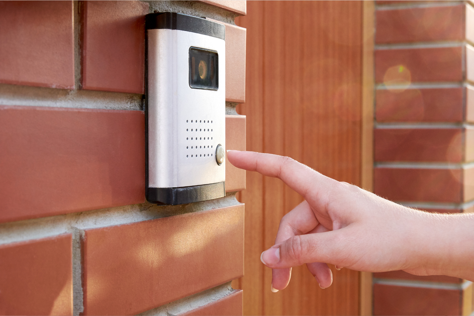 A hand pressing a button on a silver intercom with a camera on a brick wall.