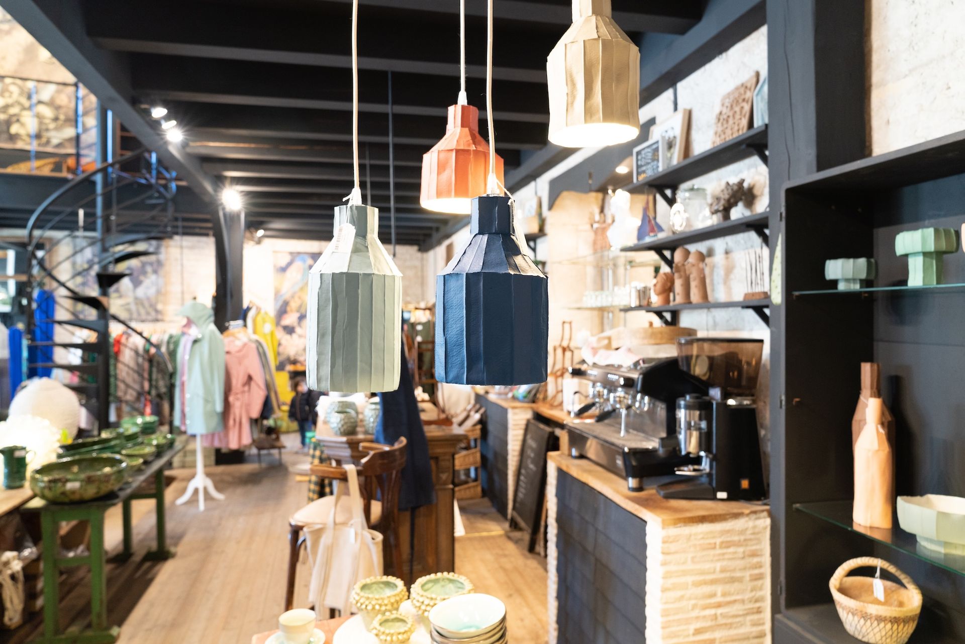 Interior shop with hanging pendant lights, clothes, shelves of items, and a spiral staircase.