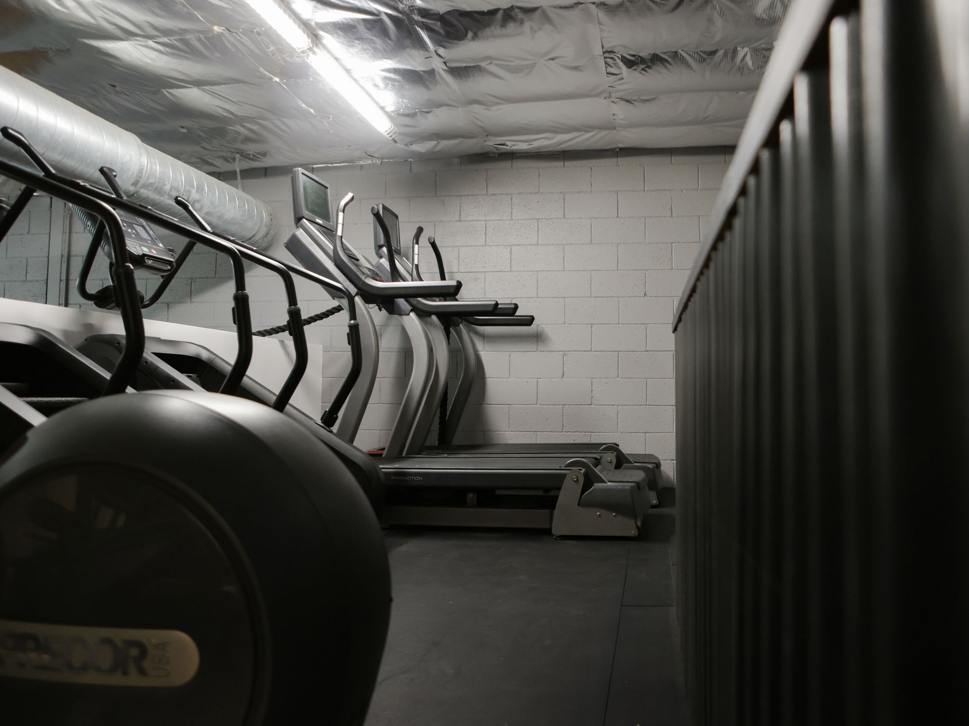 Gym with treadmills and elliptical machines, set against a white brick wall and under metallic ceiling.