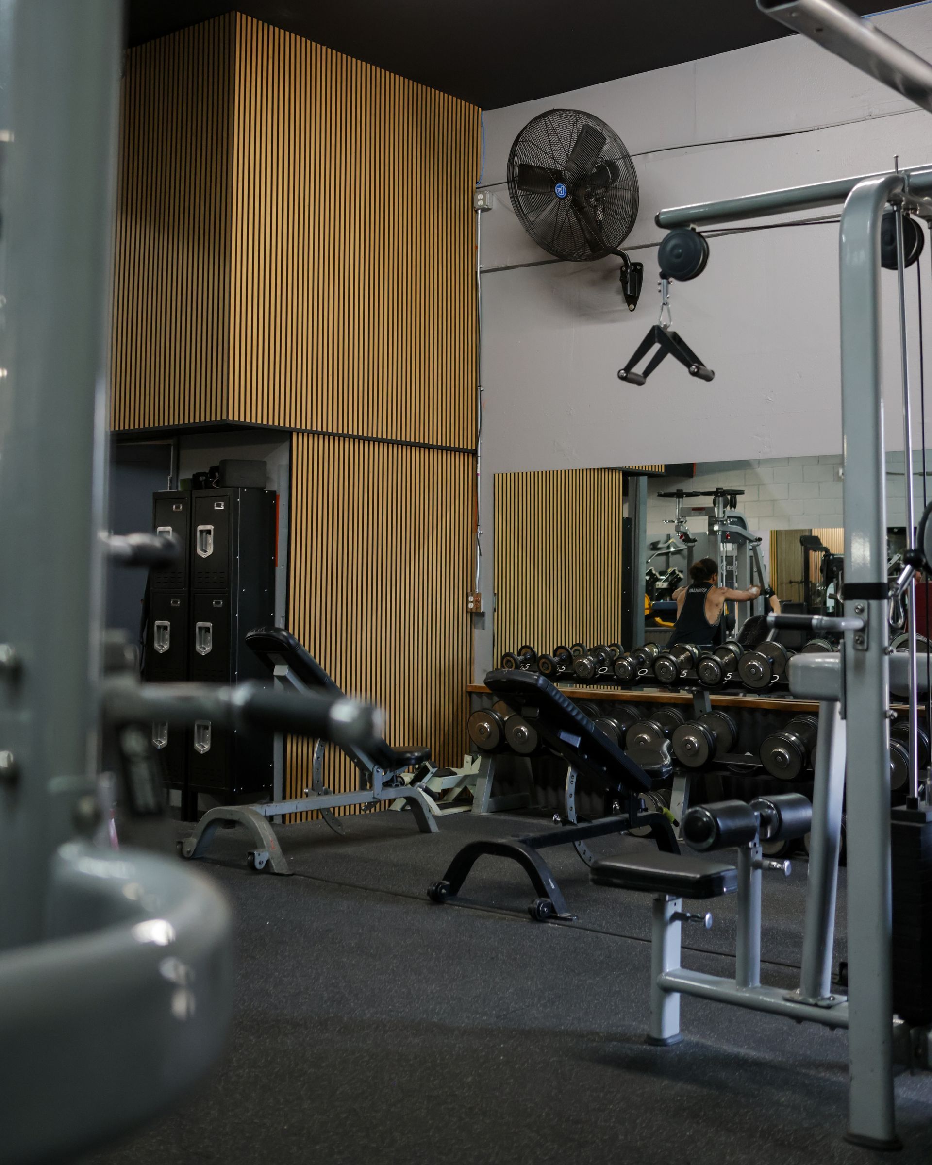Gym interior with weight machines, benches, and dumbbell racks. Dark flooring, light walls, and a large mirror.