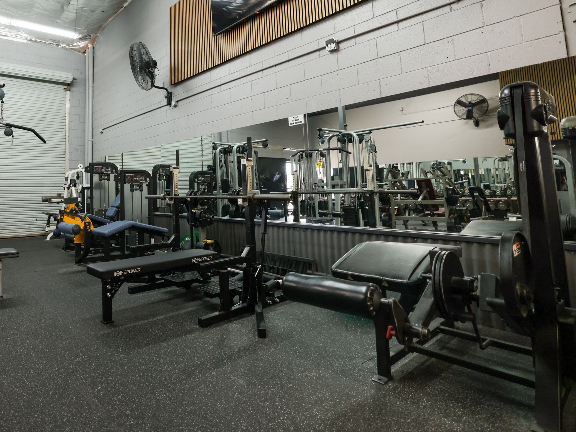 Gym interior with various weight machines, benches, and mirrors. Dark floor.