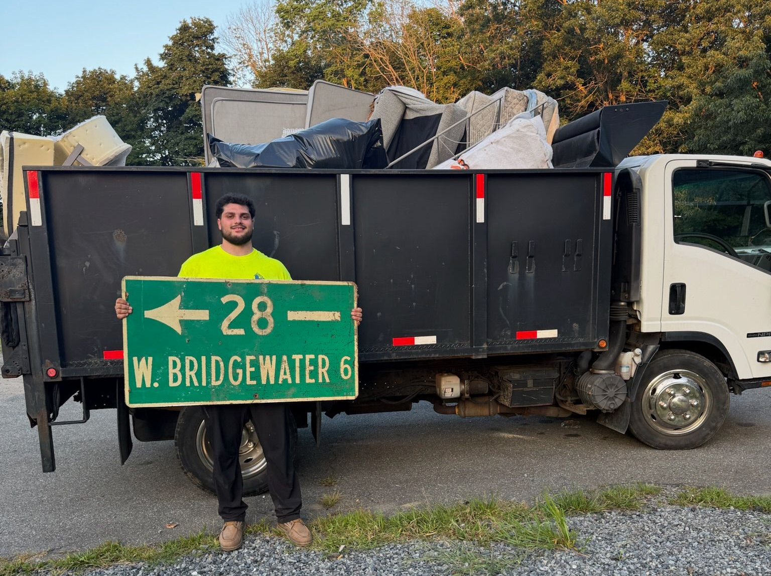 Person holding a green road sign in front of a garbage truck loaded with debris.