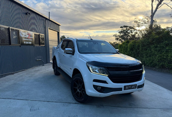 White Chevrolet Colorado pickup truck parked in front of a gray building under a cloudy sky.