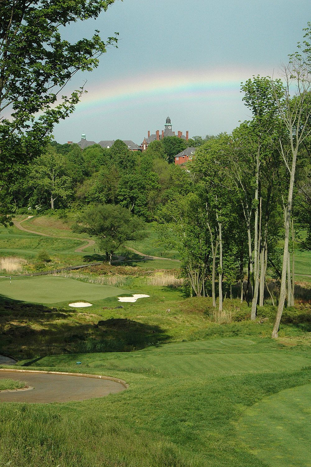 A golf course green set in a lush landscape with trees and a building in the distance under a faint rainbow.