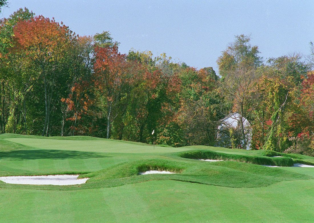A golf green with sand bunkers in the foreground and a backdrop of vibrant autumn trees under a clear blue sky.