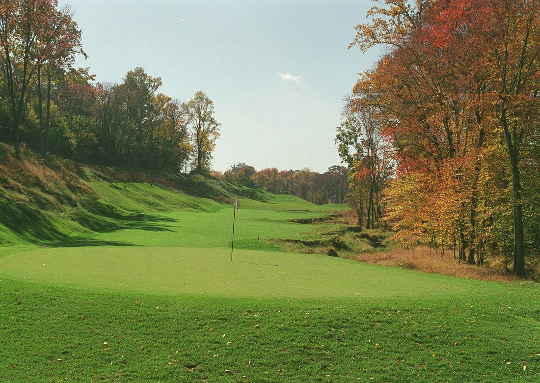 A view of a golf green in a valley, surrounded by dense trees with orange and yellow autumn foliage under a clear sky.