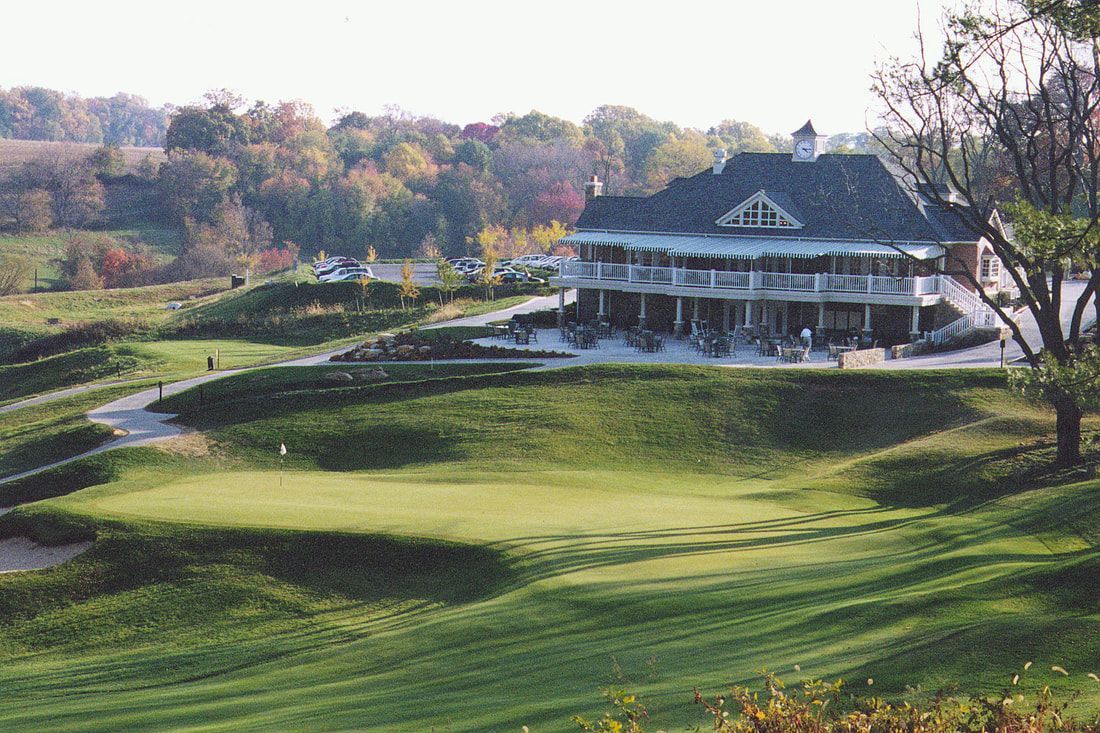 A golf course clubhouse sits on a grassy hill overlooking a putting green surrounded by trees under a clear sky.