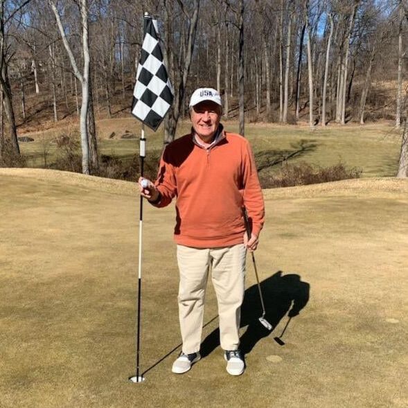 A person in an orange pullover and khaki pants stands smiling on a golf green, holding a golf ball by a checkered flag.