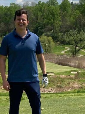 A smiling person wearing a blue polo shirt and a golf glove stands on a sunny golf course with trees in the background.