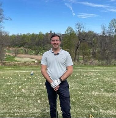 A person in a light polo shirt and dark pants stands smiling on a grassy golf course under a clear blue sky.