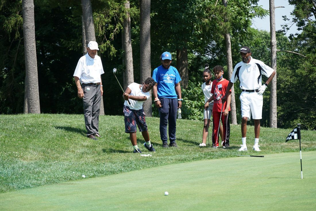 A group of people watch as a student practices a golf swing on a green, with trees in the background.