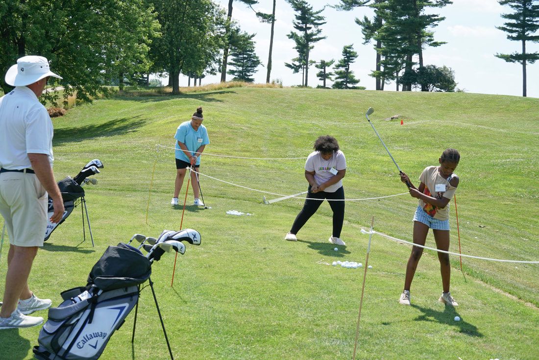 A golf instructor supervises three people practicing their golf swings on a grassy course.