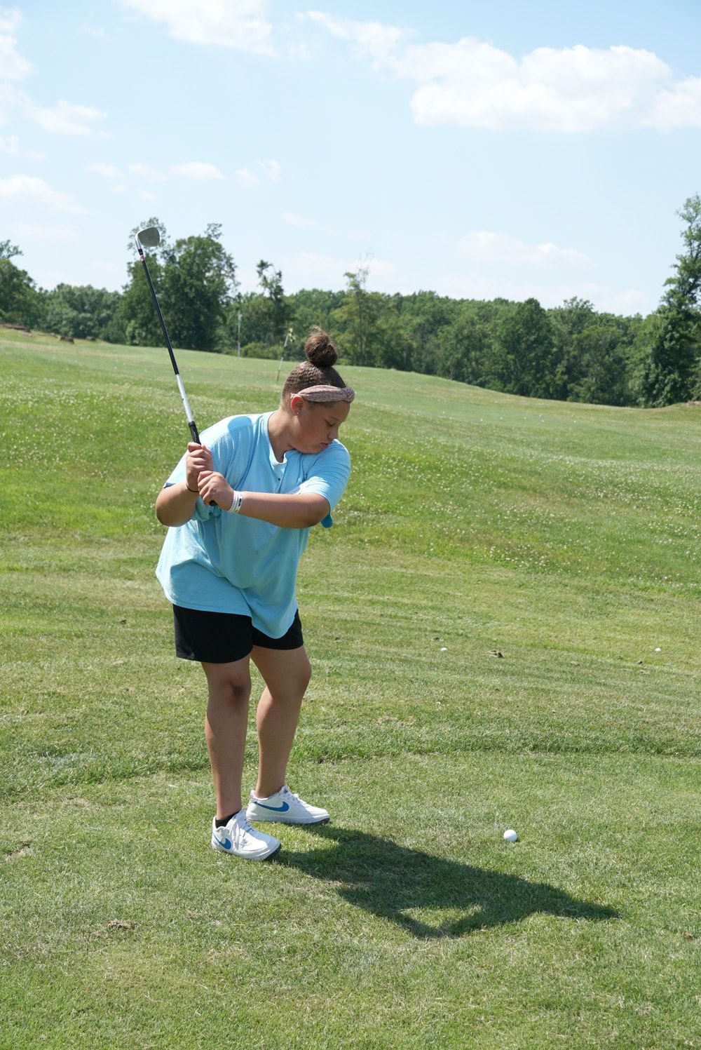 A person in a light blue shirt and black shorts swings a golf club on a grassy course under a sunny, blue sky.