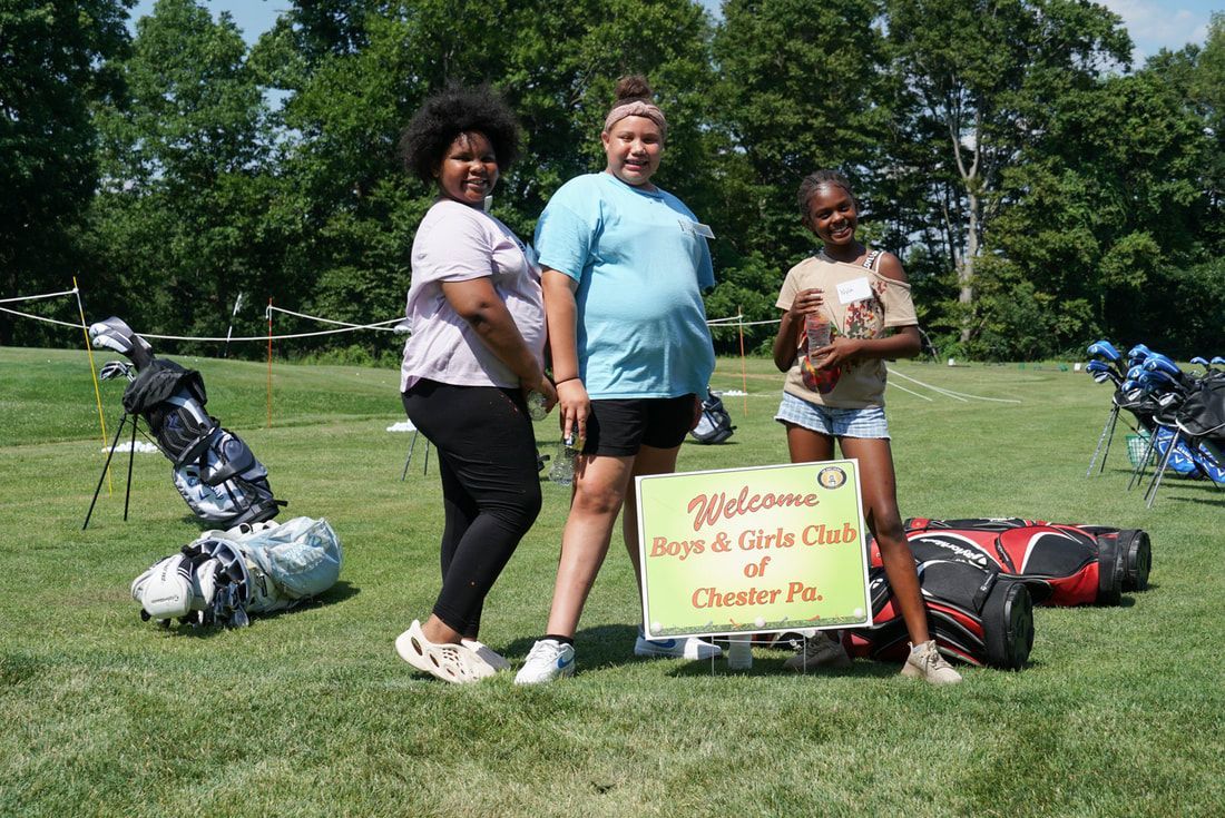 Three people stand on a grassy field with golf bags, holding a sign that says