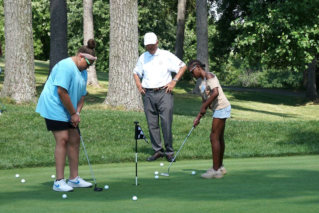 Two people putting on a golf green while an instructor observes, with numerous golf balls scattered on the grass.