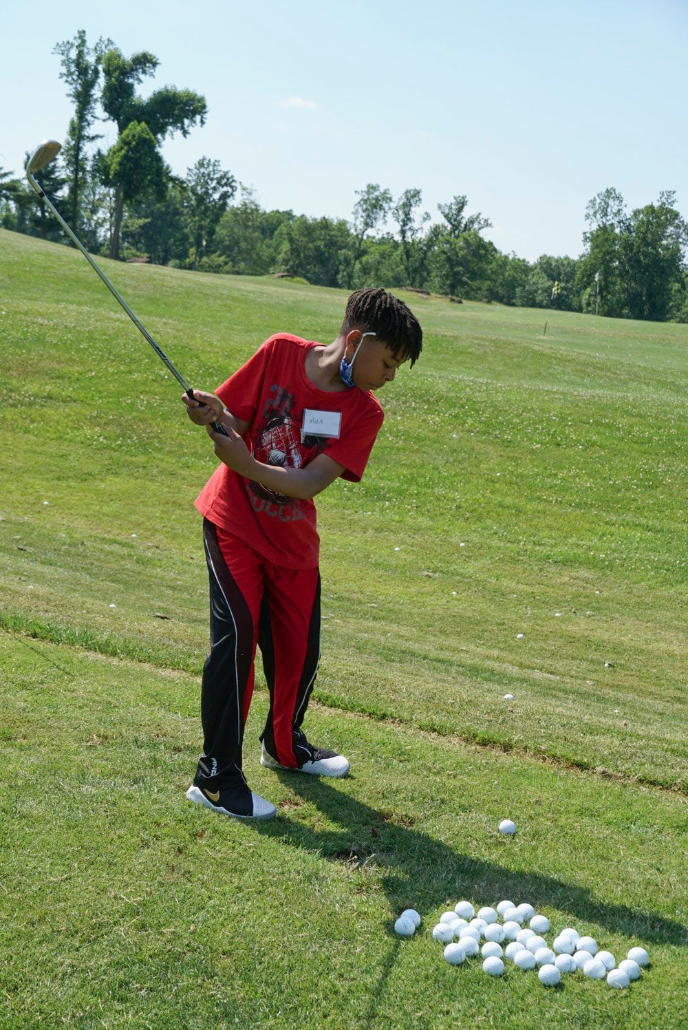 A young golfer wearing a red shirt and red and black pants swings a golf club on a grassy course near a pile of golf balls.