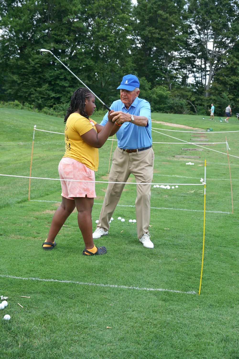 An instructor in a blue polo and hat helps a person in a yellow shirt hold a golf club on a grassy practice range.