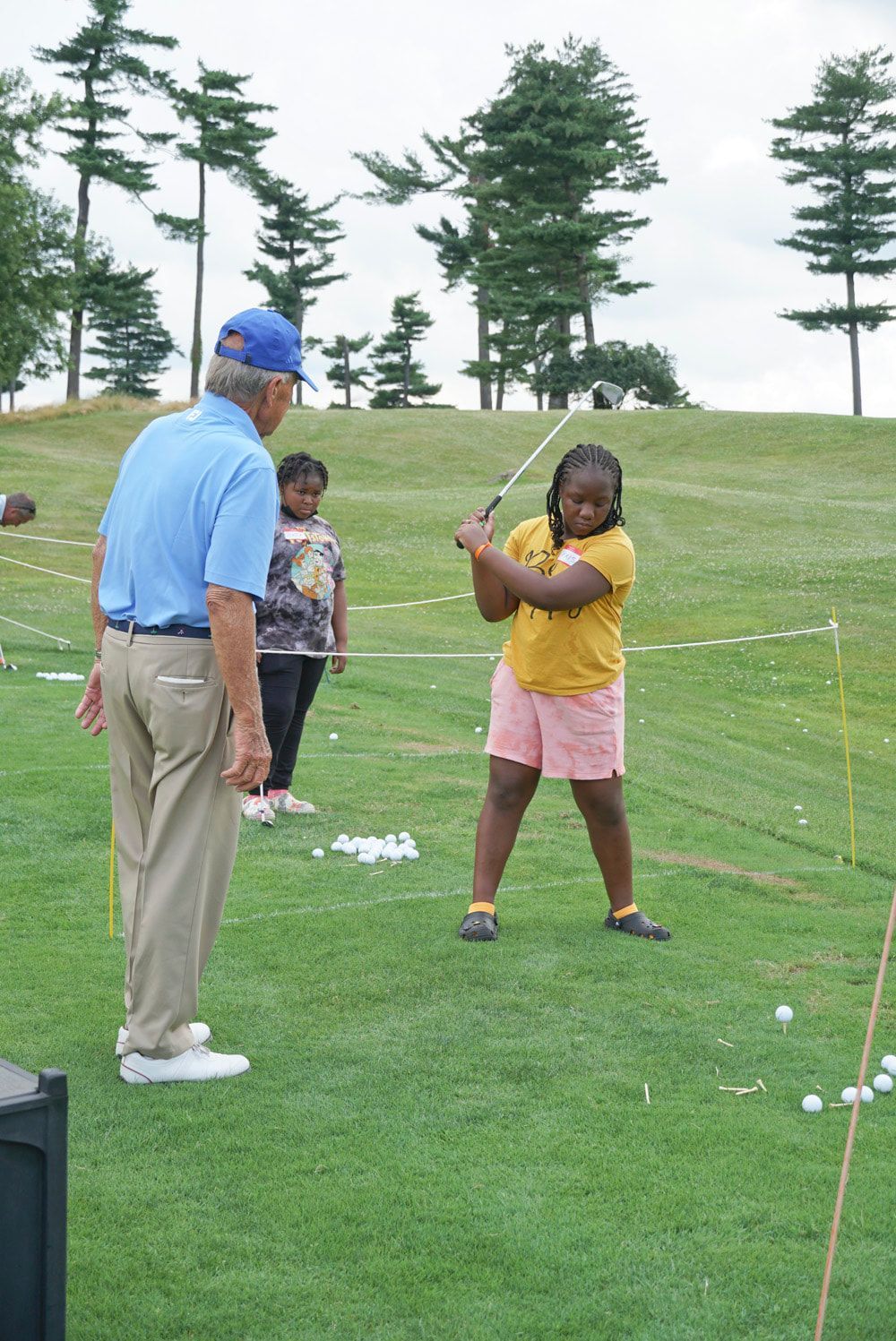 An instructor in a blue shirt watches a student practice a golf swing on a grassy outdoor course.