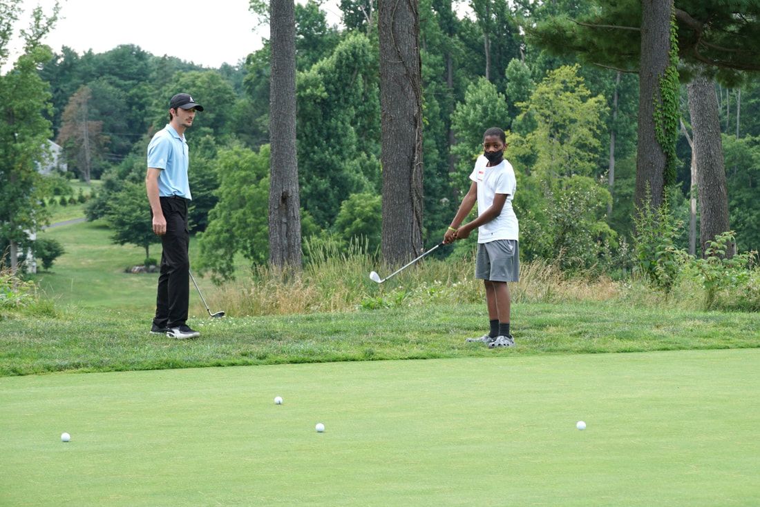 A golfer putting on a green with a coach observing in a grassy outdoor setting with trees in the background.