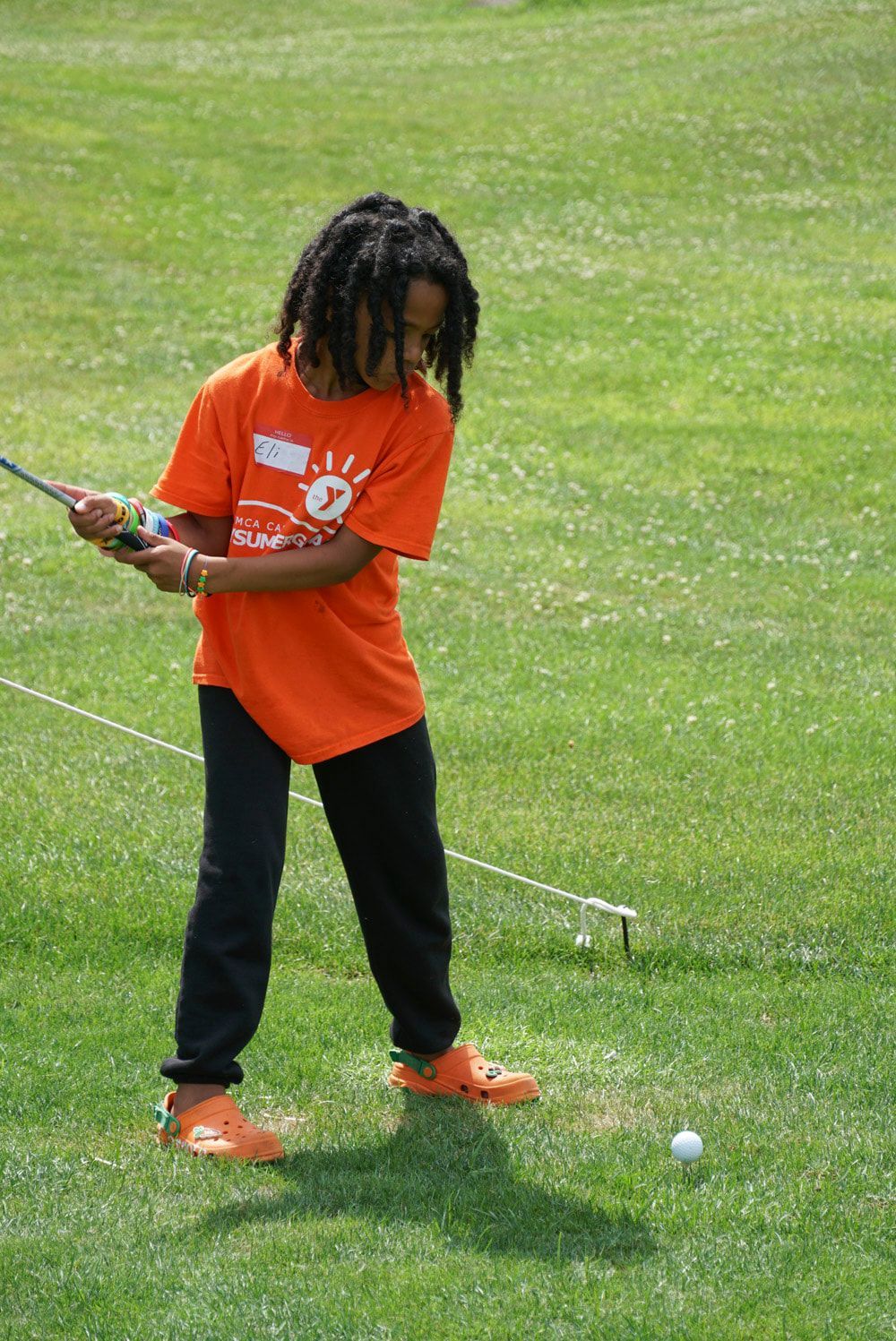 A person wearing an orange shirt and black pants stands on a grassy golf course, preparing to swing a golf club at a ball.