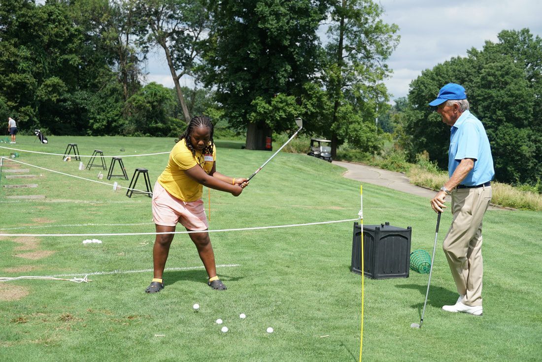 An instructor watches as a student practices a golf swing on a sunny, grassy driving range.