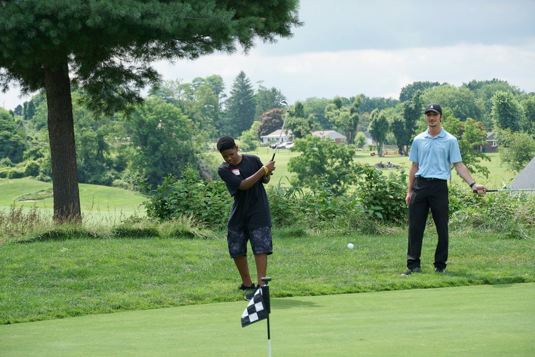 A person in a black shirt takes a golf swing near a flag on a green, with another person in a blue shirt standing nearby.