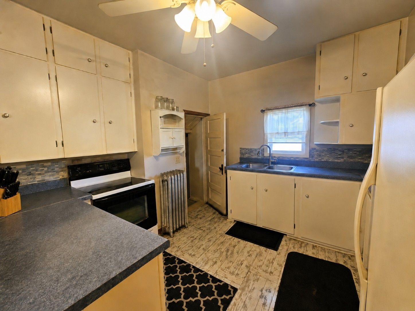 Small kitchen with cream cabinets, dark countertops, a window, and black floor mats, viewed from the corner