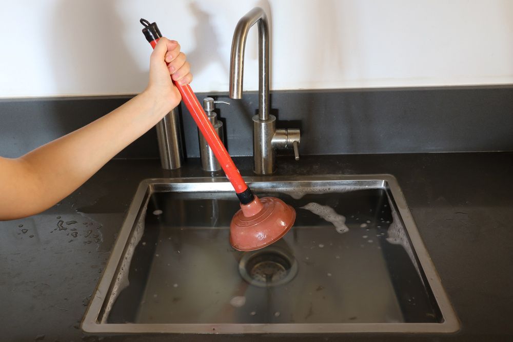 A Person Is Using A Plunger To Clean A Kitchen Sink — DMK Plumbing in Macksville, NSW