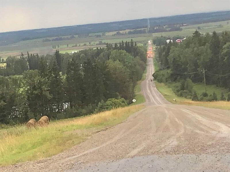 Gravel road descends a hill, trees line the sides, distant fields and hazy sky.
