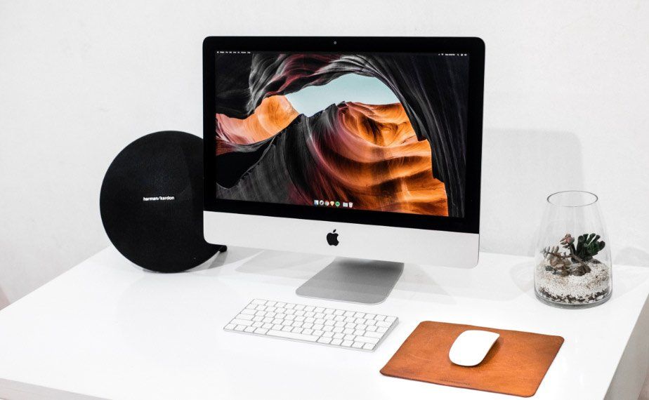 Minimal white desk with iMac, wireless keyboard, black speaker, mouse pad, and small plant.