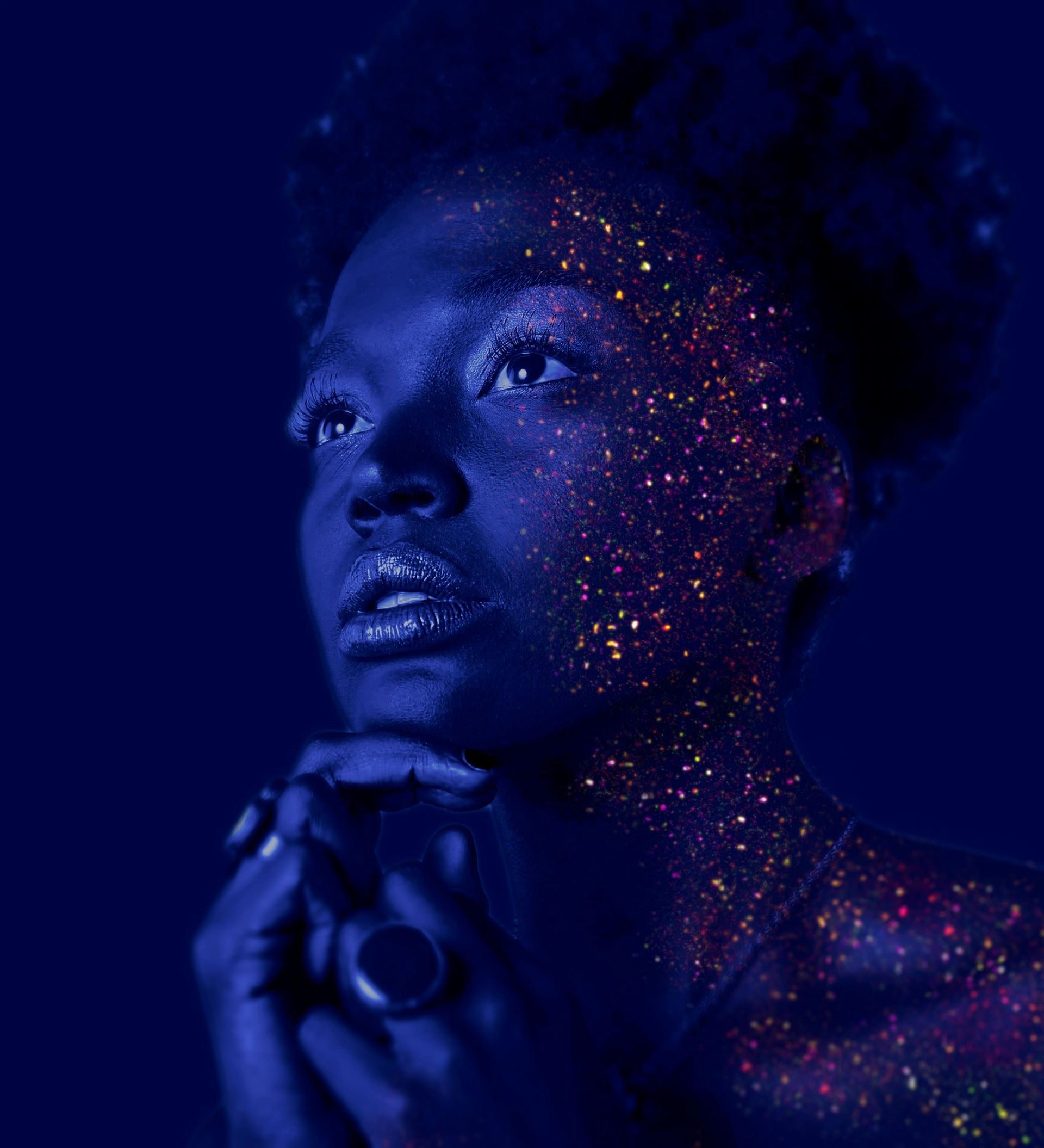 Blue-lit portrait of a person with glitter on their face and shoulders, gazing upward in thoughtful pose