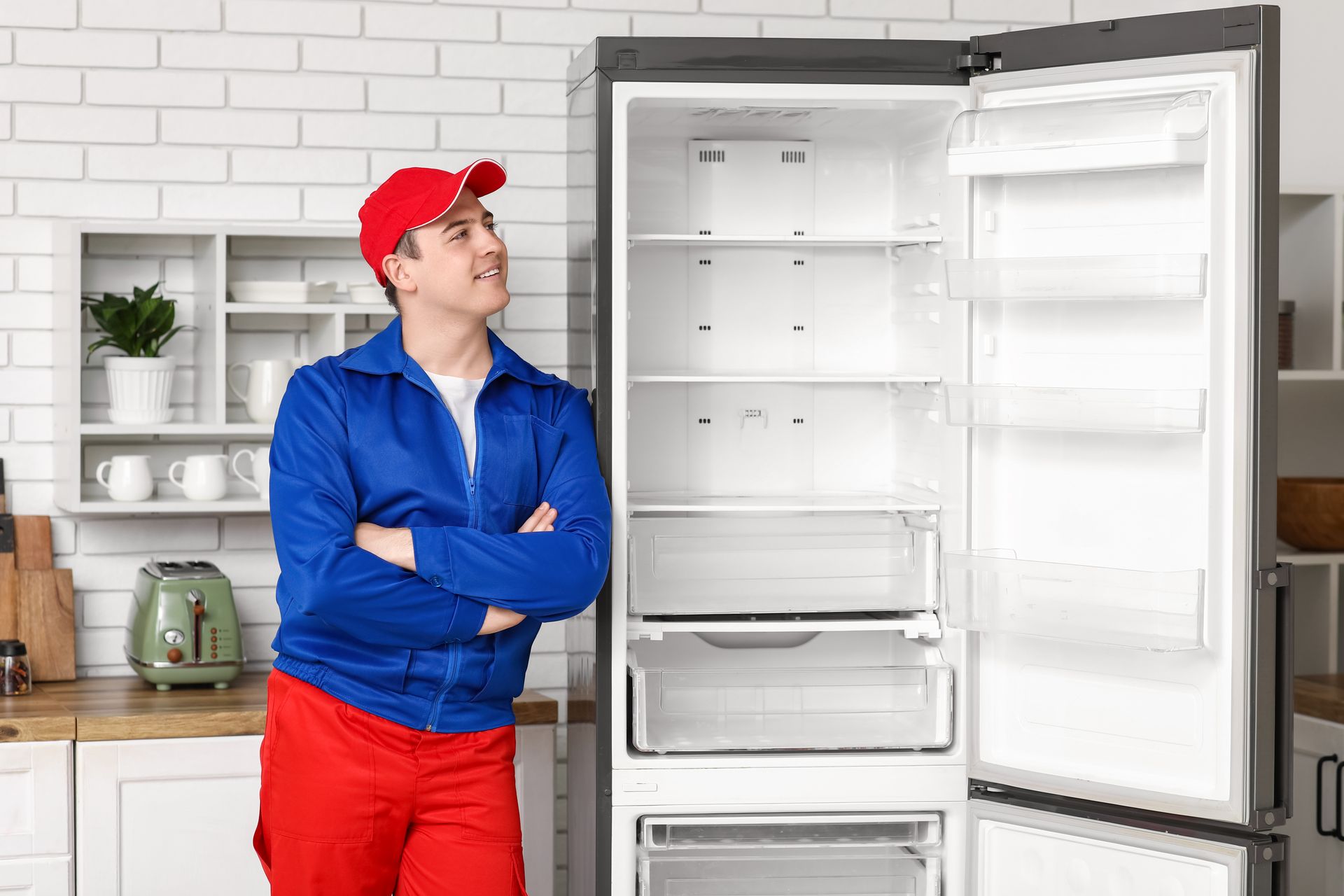 Refrigerator repairman in blue and red uniform, looking at empty fridge in kitchen.