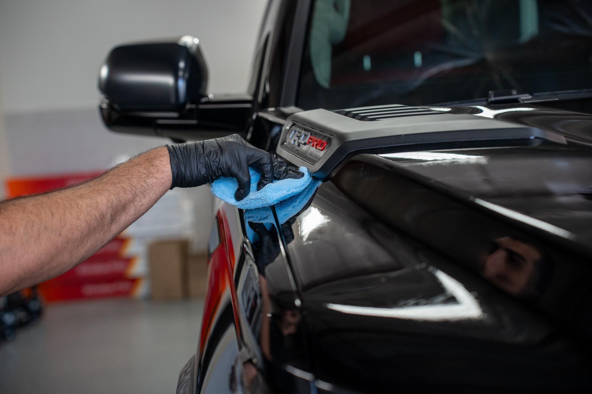 A person wearing a black glove wipes a black truck hood with a blue cloth in a garage setting.